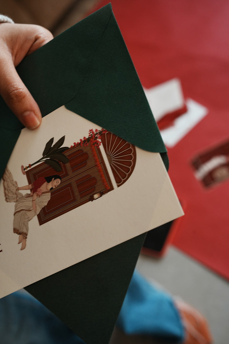 A hand holds a cream-colored art print featuring a vintage illustration of a person by an open wooden door, layered over a dark green envelope. The background is a blurred red surface.